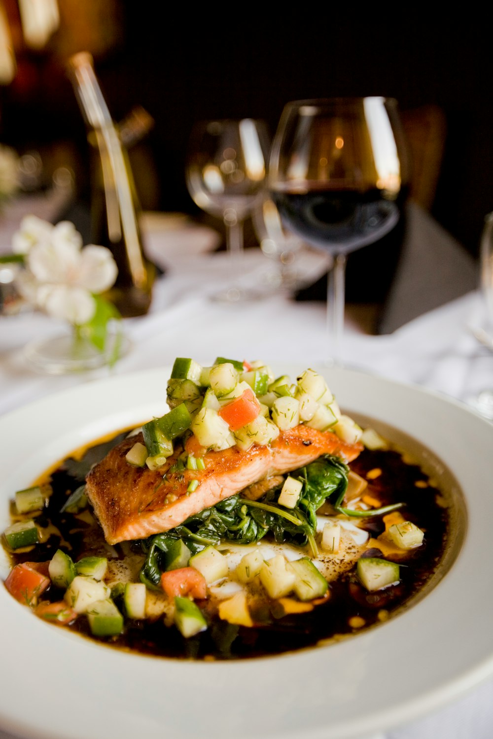 A deep dish with a meal with salmon and zucchini next to a wine glass on a white tablecloth