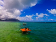 A vibrant coastal scene featuring a large expanse of calm ocean water with varying shades of blue and green. The sky is dotted with fluffy white clouds, and there is a distant view of a mountainous landform on the left side of the image. In the foreground, an orange platform with a piece of equipment on it floats on the water, accompanied by another distant platform.