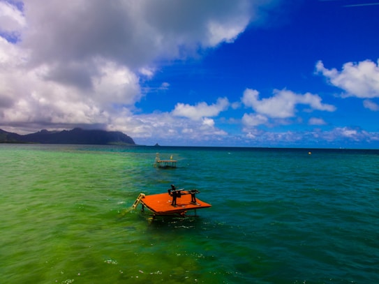 A vibrant coastal scene featuring a large expanse of calm ocean water with varying shades of blue and green. The sky is dotted with fluffy white clouds, and there is a distant view of a mountainous landform on the left side of the image. In the foreground, an orange platform with a piece of equipment on it floats on the water, accompanied by another distant platform.