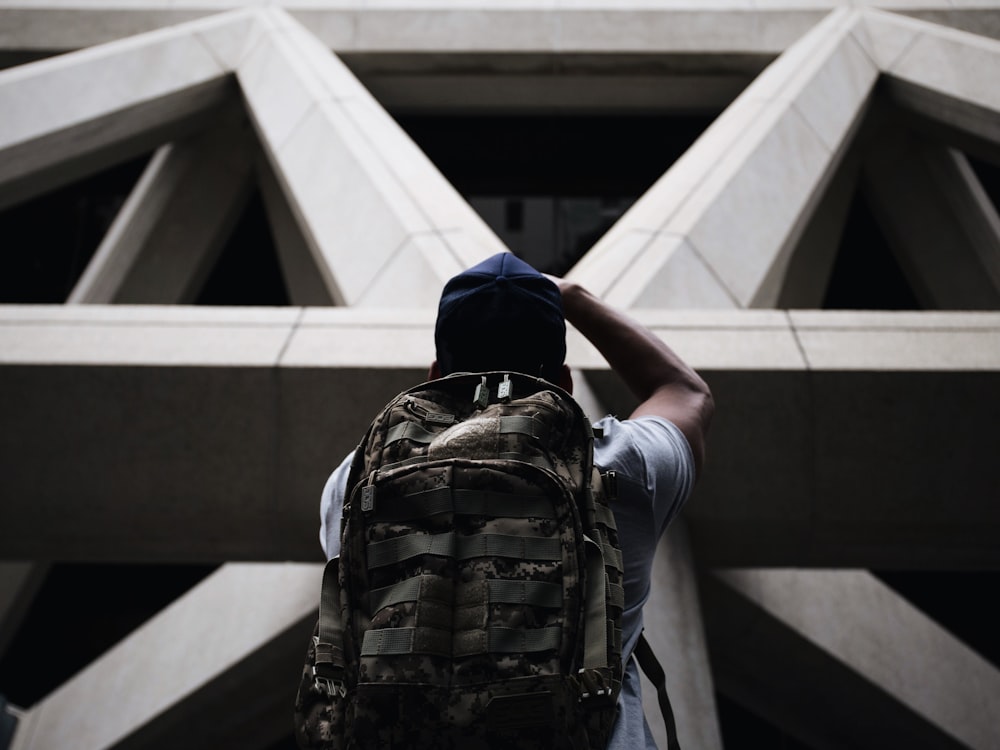 person wearing shirt and hiking bag looking in front of grey concrete structure while standing during daytime