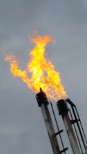 Bright orange flames are rising from the top of metal flare stacks against a cloudy gray sky.