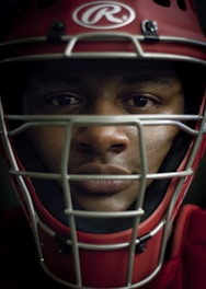A close-up of a person wearing a red sports helmet with a metallic face guard. The logo on the helmet is visible, and the person's expression appears focused and intense.