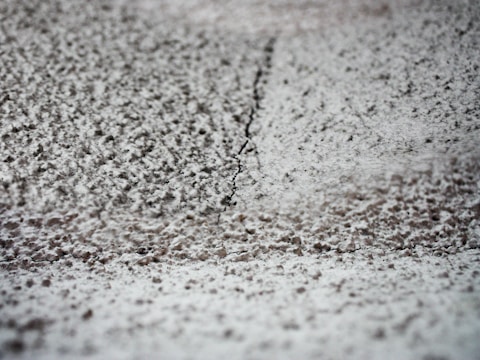 A close-up view of a textured surface with a thin crack running vertically through the middle. The surface appears to be rough and uneven, with small, granular particles spread across it.