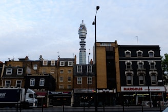 A cityscape featuring a mix of old and modern architecture. In the foreground, a row of traditional brick buildings with various businesses, including a restaurant and shoe store, line the street. A large telecommunications tower rises in the background, dominating the skyline. A truck is parked on the street, and a lamppost stands in the middle of the scene.