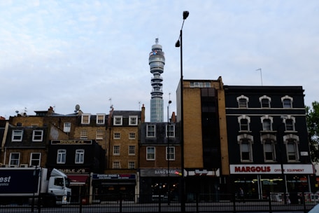 A cityscape featuring a mix of old and modern architecture. In the foreground, a row of traditional brick buildings with various businesses, including a restaurant and shoe store, line the street. A large telecommunications tower rises in the background, dominating the skyline. A truck is parked on the street, and a lamppost stands in the middle of the scene.