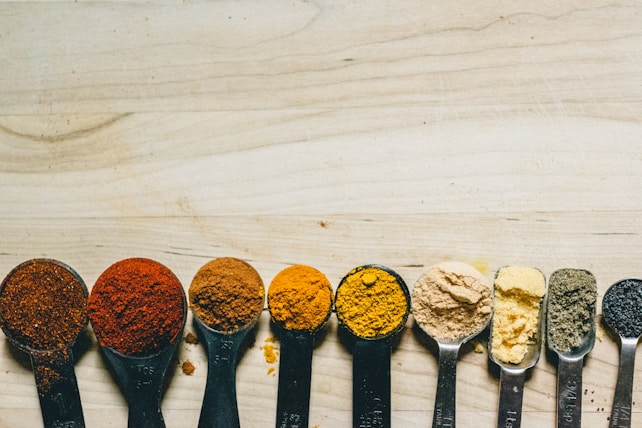 A series of measuring spoons are aligned horizontally on a wooden surface, each filled with a different type of spice or powdered ingredient. The colors range from deep reds and oranges to yellows, light browns, and blacks. The spoons are made of metal and are evenly spaced.