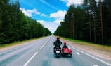 Two people ride a motorcycle with a sidecar down an empty, straight road surrounded by dense green forest. The sky is bright blue with fluffy white clouds, creating a sense of open space and freedom. The road stretches off into the horizon, emphasizing the journey ahead.
