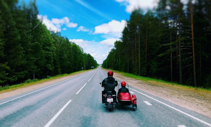 Two people ride a motorcycle with a sidecar down an empty, straight road surrounded by dense green forest. The sky is bright blue with fluffy white clouds, creating a sense of open space and freedom. The road stretches off into the horizon, emphasizing the journey ahead.