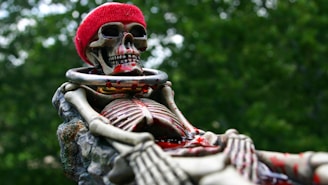 A skeleton is adorned with a red hat and a metallic collar, sitting against a background of blurred greenery. The skeleton appears detailed, with visible bones and red paint accentuating certain areas. The scene conveys a mix of eeriness and whimsy.