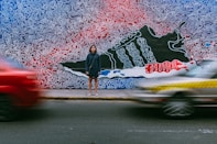time lapse photography of woman wearing blue jacket standing on concrete pathway in front of two vehicles passing on concrete road during daytime