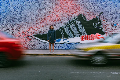 time lapse photography of woman wearing blue jacket standing on concrete pathway in front of two vehicles passing on concrete road during daytime