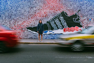 time lapse photography of woman wearing blue jacket standing on concrete pathway in front of two vehicles passing on concrete road during daytime