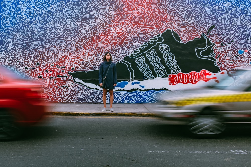 time lapse photography of woman wearing blue jacket standing on concrete pathway in front of two vehicles passing on concrete road during daytime