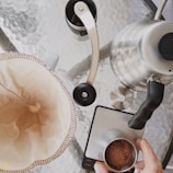 A top-down view of coffee-making equipment on a glass table, including a stainless steel kettle, a coffee grinder, a scale, and a coffee filter holder. A person's hand is adding ground coffee to the filter, preparing for a pour-over coffee brewing process.