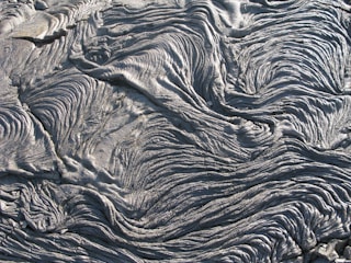 An intricate pattern of hardened lava flows, displaying a series of wavy, rippling textures resembling folds of fabric or waves. The surface appears rough and slightly glossy, with a predominantly grayish color.