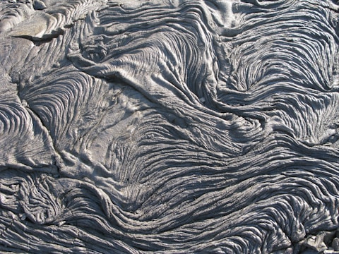 An intricate pattern of hardened lava flows, displaying a series of wavy, rippling textures resembling folds of fabric or waves. The surface appears rough and slightly glossy, with a predominantly grayish color.