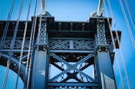 A detailed view of a metal bridge structure, showcasing intricate cross-bracing and supporting cables against a clear blue sky. The metalwork features a pattern of bolts and rivets, emphasizing industrial design.