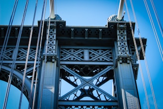 A detailed view of a metal bridge structure, showcasing intricate cross-bracing and supporting cables against a clear blue sky. The metalwork features a pattern of bolts and rivets, emphasizing industrial design.