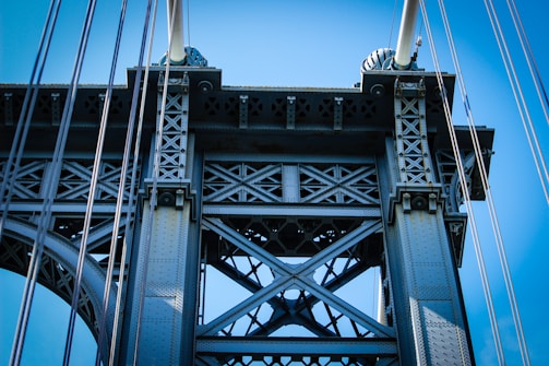 A detailed view of a metal bridge structure, showcasing intricate cross-bracing and supporting cables against a clear blue sky. The metalwork features a pattern of bolts and rivets, emphasizing industrial design.