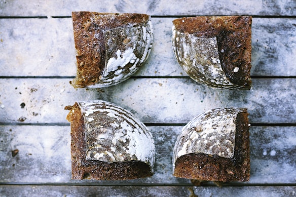 Four pieces of rustic, crusty bread are arranged on a weathered wooden surface. The bread has a dark, textured crust with a dusting of flour, suggesting a handmade, artisanal quality. The wood background has a grayish tone with visible grain and imperfections, enhancing the rustic feel.