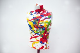 A mannequin torso is covered in an abstract pattern of vibrant splashes and streaks of paint in various colors, set against a plain background.