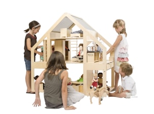 A group of children are engaged in play around a large, wooden dollhouse. The dollhouse is well-crafted with various rooms and pieces of furniture. Dolls are placed throughout, and the children appear to be interacting with the dolls and the house. The children are focused and seem to be enjoying their imaginative play. The scene is well-lit and the background is plain, enhancing the focus on the children and the dollhouse.