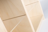The image features an angled view of a light-colored wooden shelf or organizer. The wood grain is visible, giving a natural and minimalist aesthetic. The design includes slots or dividers, suggesting functionality for storage or organization.