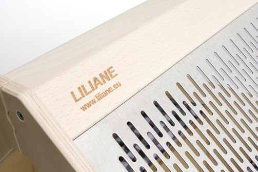 The image features a wooden structure with a carved name and website link engraved on the surface. Adjacent to the wooden area is a metallic section with evenly spaced vertical slits, providing a sleek and modern design.