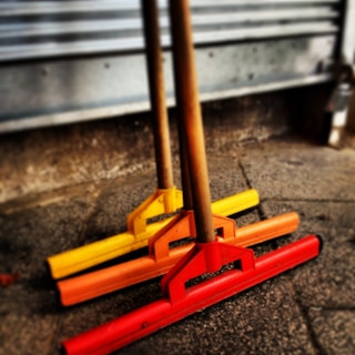 Three push brooms with wooden handles, arranged side by side, have bright red, orange, and yellow heads. They rest against a textured, concrete surface in front of a metallic, corrugated backdrop.