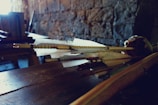 Several arrows with detailed fletching are resting on a wooden surface, with a rustic stone wall in the background. The lighting is dim, creating a moody atmosphere.
