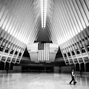 A large indoor space featuring modern architectural design with tall, curved rib-like structures lining the walls. The ceiling has a narrow skylight, and an American flag is prominently displayed on a central platform. A person is pushing a cleaning cart across the smooth, reflective floor.