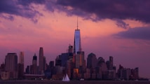 The image captures a city skyline at sunset with a prominent skyscraper. The sky is filled with purple and pink hues, casting a warm glow over the buildings. Clouds are scattered across the sky, adding texture. Below, a sailboat can be seen on the water in the foreground.