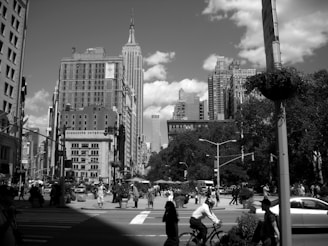 A bustling city street scene features tall skyscrapers, including an iconic building in the background. The foreground shows a busy intersection with pedestrians walking and a person riding a bicycle. Trees and streetlights line the street, adding to the urban landscape. The photo is in black and white, giving it a classic, timeless feel.