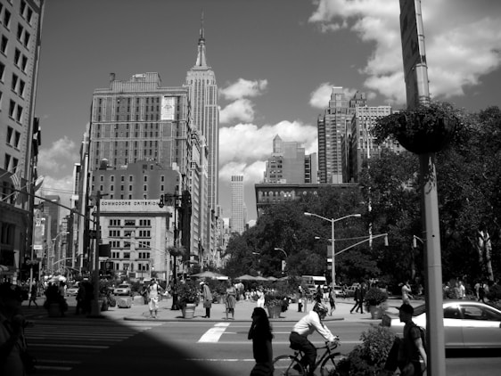 A bustling city street scene features tall skyscrapers, including an iconic building in the background. The foreground shows a busy intersection with pedestrians walking and a person riding a bicycle. Trees and streetlights line the street, adding to the urban landscape. The photo is in black and white, giving it a classic, timeless feel.
