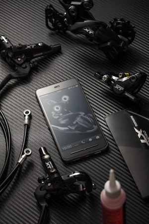 A flat lay arrangement featuring bicycle components, including Shimano XT series disc brakes and cables. A smartphone displays a matching bicycle part image on its screen, creating a cohesive theme. The background is a textured, dark carbon fiber-like surface, enhancing the sleek look of the components.