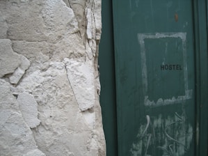 A weathered wall with peeling plaster stands next to a green door with the word 'HOSTEL' written on it. The door shows signs of wear, with scuff marks and faded paint, adding to the overall aged appearance of the scene.
