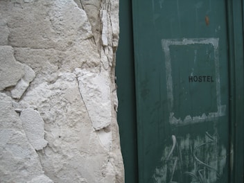 A weathered wall with peeling plaster stands next to a green door with the word 'HOSTEL' written on it. The door shows signs of wear, with scuff marks and faded paint, adding to the overall aged appearance of the scene.