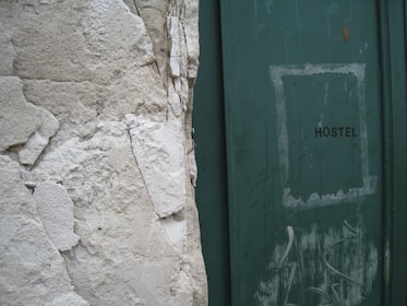 A weathered wall with peeling plaster stands next to a green door with the word 'HOSTEL' written on it. The door shows signs of wear, with scuff marks and faded paint, adding to the overall aged appearance of the scene.