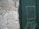 A weathered wall with peeling plaster stands next to a green door with the word 'HOSTEL' written on it. The door shows signs of wear, with scuff marks and faded paint, adding to the overall aged appearance of the scene.