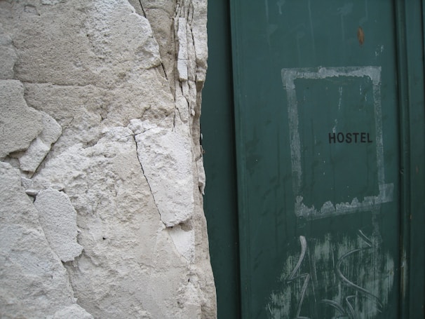 A weathered wall with peeling plaster stands next to a green door with the word 'HOSTEL' written on it. The door shows signs of wear, with scuff marks and faded paint, adding to the overall aged appearance of the scene.