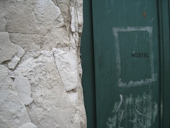 A weathered wall with peeling plaster stands next to a green door with the word 'HOSTEL' written on it. The door shows signs of wear, with scuff marks and faded paint, adding to the overall aged appearance of the scene.