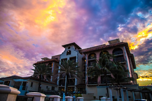 A large residential building with Mediterranean architectural features is surrounded by palm trees. The colorful sky at sunset creates a dramatic backdrop with hues of orange, yellow, pink, and blue.