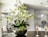 A modern, open-plan kitchen and dining area featuring a prominent orchid plant in the foreground, with delicate white and green flowers. There are two wine glasses filled with rosé on a dark round table. The kitchen has white cabinetry, a double-door refrigerator, and pendant lights. In the background, a dining table is set with chairs and another flower arrangement.