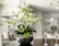 A modern, open-plan kitchen and dining area featuring a prominent orchid plant in the foreground, with delicate white and green flowers. There are two wine glasses filled with rosé on a dark round table. The kitchen has white cabinetry, a double-door refrigerator, and pendant lights. In the background, a dining table is set with chairs and another flower arrangement.