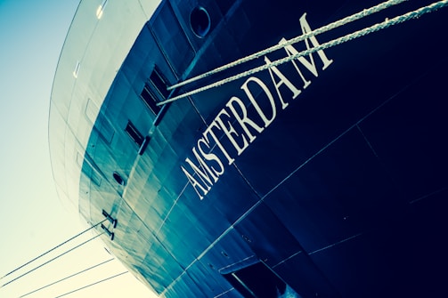 The image captures the side view of a ship with the word 'AMSTERDAM' prominently displayed. The ship is painted in dark tones with a sleek, modern design. Two thick ropes run across the foreground, adding depth and interest.