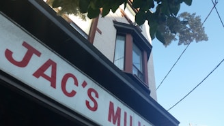 A building with a sign that partially reads 'JAC'S' in red letters on a white background. The structure features a white brick exterior with a bay window framed in dark brown. Overhead, green tree leaves partially obscure the view, and electrical wires are visible against a clear blue sky.