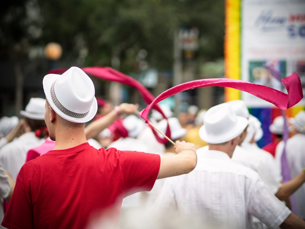 selective focus photography of man wearing pink t-shirt and white fedora hat holding pink ribbon with stick waving on the air during daytime