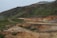 A rugged, mountainous landscape with green and brown hues features a hiking trail winding through it. Small groups of people walking along the path can be seen, indicating a sense of exploration and adventure. The terrain appears natural and slightly barren, with subtle steam rising in the background.