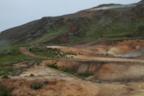 A rugged, mountainous landscape with green and brown hues features a hiking trail winding through it. Small groups of people walking along the path can be seen, indicating a sense of exploration and adventure. The terrain appears natural and slightly barren, with subtle steam rising in the background.