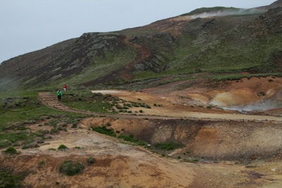 A rugged, mountainous landscape with green and brown hues features a hiking trail winding through it. Small groups of people walking along the path can be seen, indicating a sense of exploration and adventure. The terrain appears natural and slightly barren, with subtle steam rising in the background.